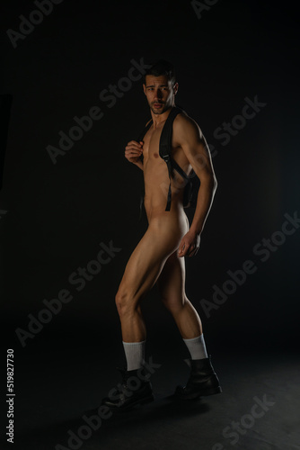 Sexy nude man posing in a dark studio in black boots with a black backpack