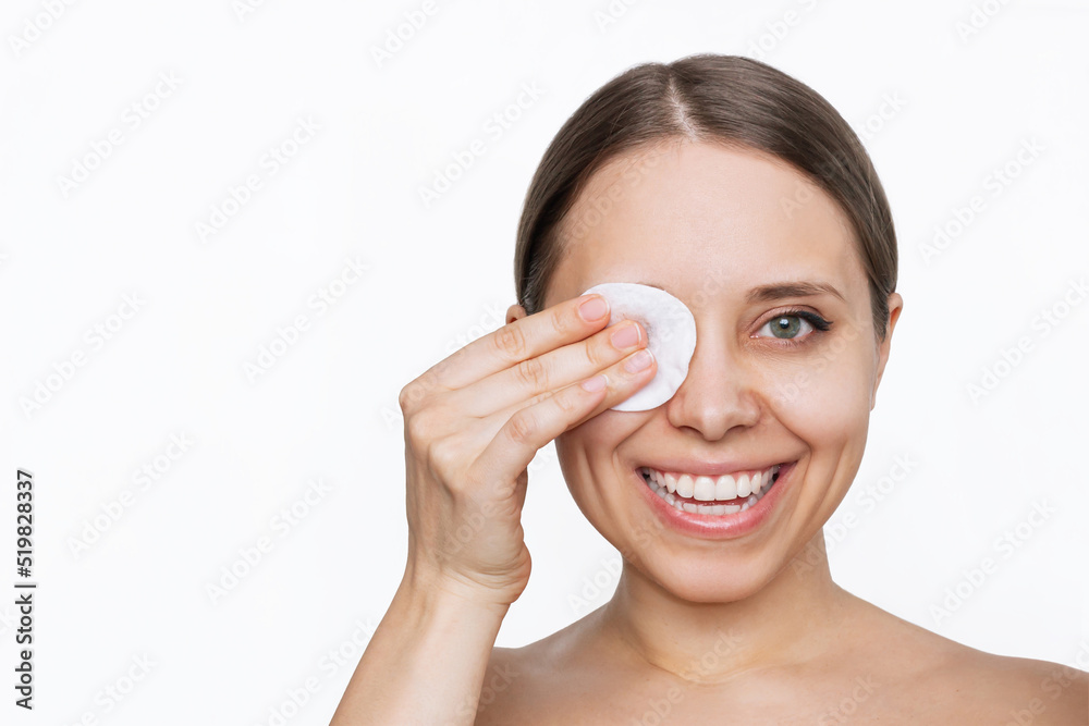 Close up of a young caucasian smiling woman cleaning her face with cotton pad removing makeup from the eyes isolated on a white background. Skin care, cosmetology. The girl washes off her makeup Close up of a young caucasian smiling woman cleaning her face with cotton pad removing makeup from the eyes isolated on a white background. Skin care, cosmetology. The girl washes off her makeup