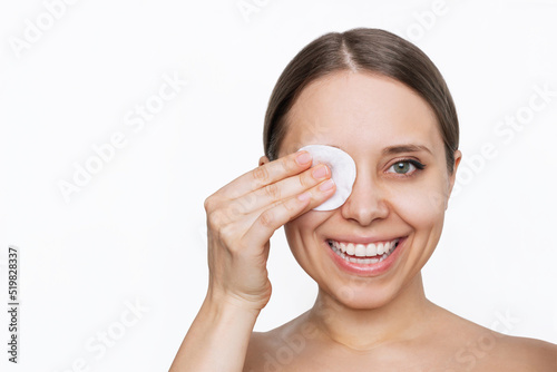 Close up of a young caucasian smiling woman cleaning her face with cotton pad removing makeup from the eyes isolated on a white background. Skin care, cosmetology. The girl washes off her makeup