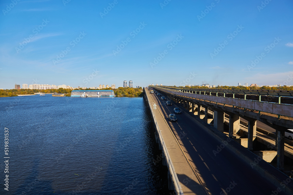 Autumn of the cityscape of Kyiv overlooking the Dnipro River, the railway line and the metro bridge, Hydropark and the left bank of the Rusanovka district under bright sunlight.