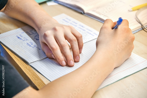 Schoolboy in black t-shirt does homework sitting at wooden desk at home. Hand of boy holds pen writing text in exercise book close view