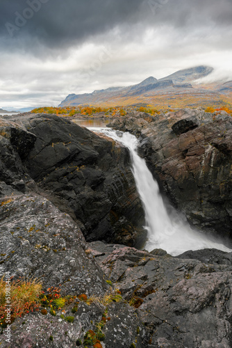 Mighty scenic waterfall under dramatic sky. Stuor Muorkke waterfall in Stora Sjofallet national park, Sweden. Adventure in arctic wilderness. Autumn colors in the arctic.