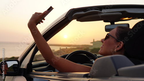 Attractive woman makes selfie and takes photos of the sunset from her convertible car parked in front of the beach, enjoying the sunset alone and relaxing with the view