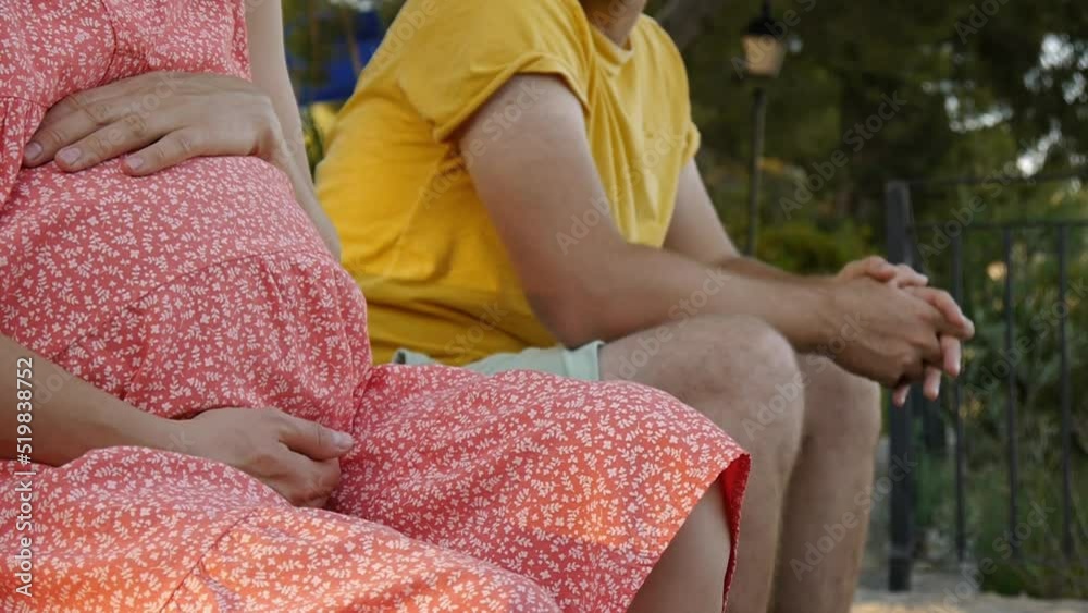 Pregnant young caucasian woman touching her belly. Couple spending time together sitting on the bench in the park.