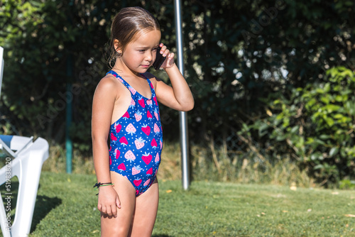 Little girl in a swimsuit, standing on the edge of the pool, talking on the mobile phone. Childhood, children, smartphone, technology, vacation, internet and fun concept.