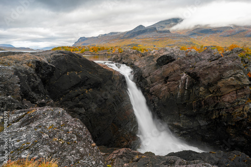 Mighty scenic waterfall under dramatic sky. Stuor Muorkke waterfall in Stora Sjofallet national park, Sweden. Adventure in arctic wilderness. Autumn colors in the arctic.