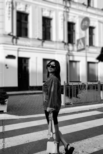Beautiful girl with sunglasses. A model at a pedestrian crossing. Stylish young woman in a black dress and jeans jacket. Street style shooting. Women's fashion. Black and white photo