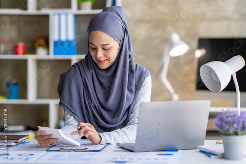 Attractive Muslim woman wearing a hijab working in a business office Use a calculator and laptop for calculating money on a wooden desk in the office. Taxes, accounting, financial concepts.