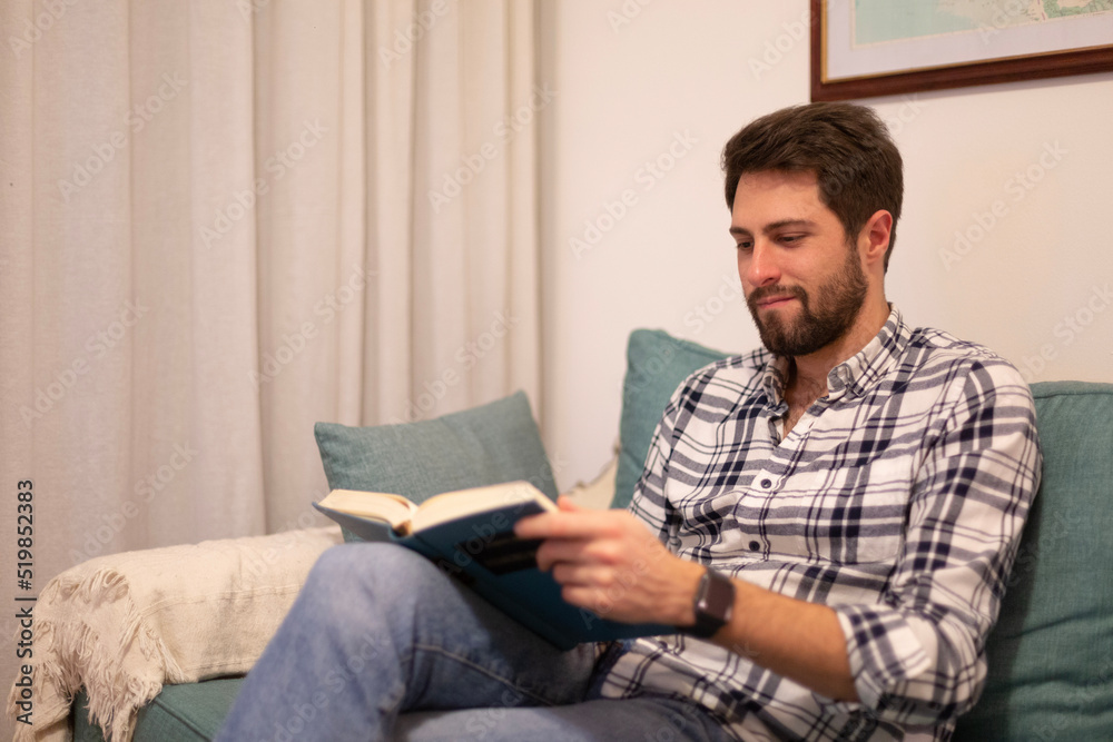 Portrait handsome bearded man wearing squared shirt. Young man sitting in a couch reading book and relaxing. Horizontal.