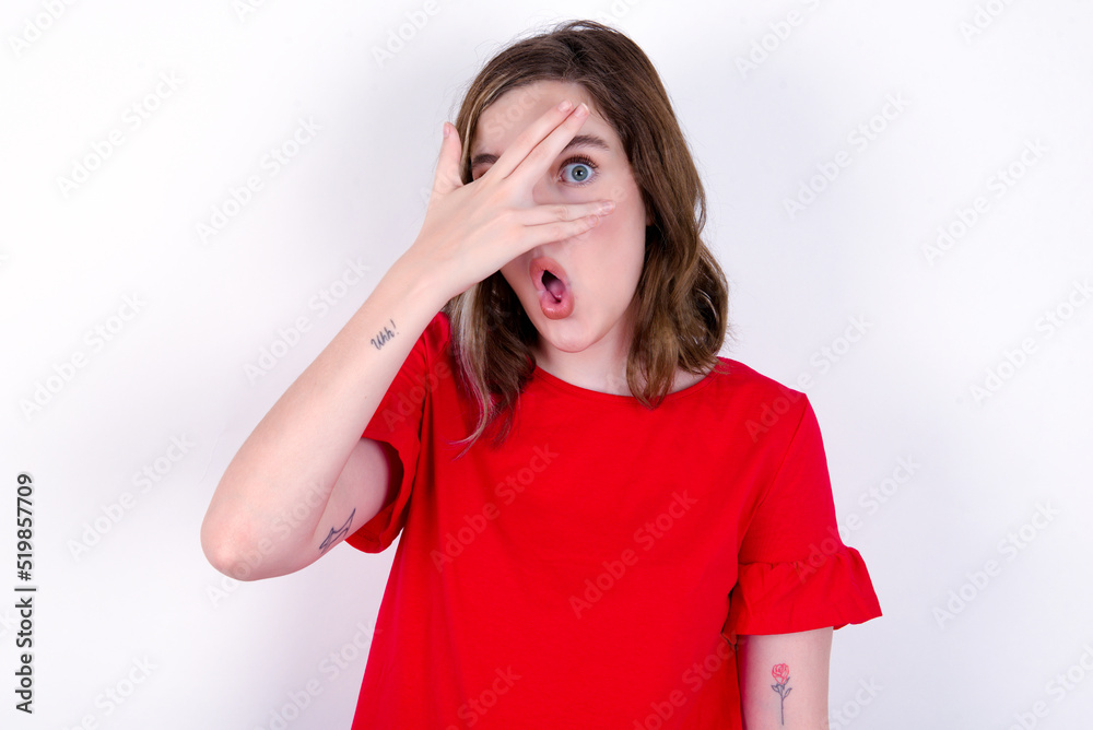 young caucasian woman wearing red T-shirt over white background peeking in shock covering face and eyes with hand, looking through fingers with embarrassed expression. young caucasian woman wearing red T-shirt over white background peeking in shock covering face and eyes with hand, looking through fingers with embarrassed expression.