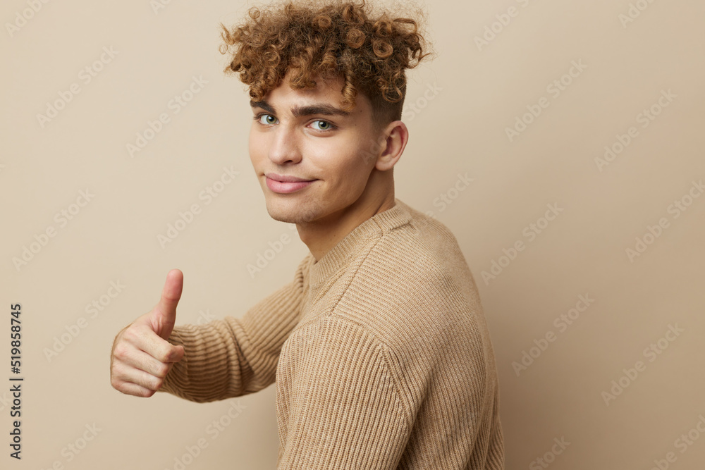 a close horizontal portrait of a handsome, charming man with curly hair standing in a beige sweater on a beige background, smiling pleasantly at the camera showing a thumbs up a close horizontal portrait of a handsome, charming man with curly hair standing in a beige sweater on a beige background, smiling pleasantly at the camera showing a thumbs up