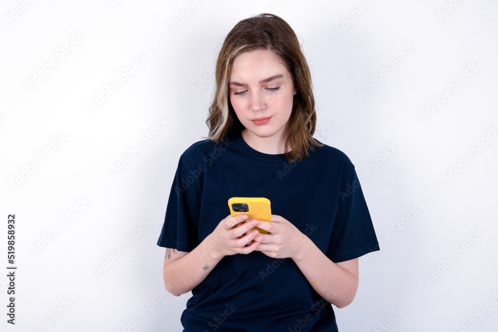 Excited young caucasian woman wearing black T-shirt over white background winking and eye hold smart phone use read social network news Excited young caucasian woman wearing black T-shirt over white background winking and eye hold smart phone use read social network news