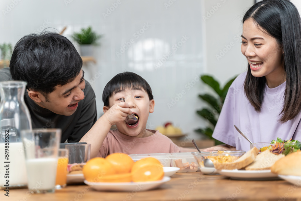 Happy Asian family enjoying breakfast together on dinning table. Young Asian mother feeding vegetable to her son while having breakfast in the kitchen. Healthy food family activity lifestyle concept.