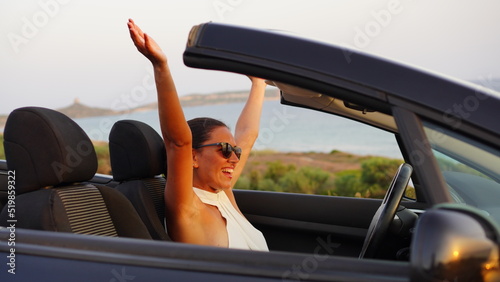 Lonely woman in a black convertible car looks at the sunset in front of the sea and is moved by how wonderful it is and raises her arms with a feeling of freedom and happiness during her vacation alon photo