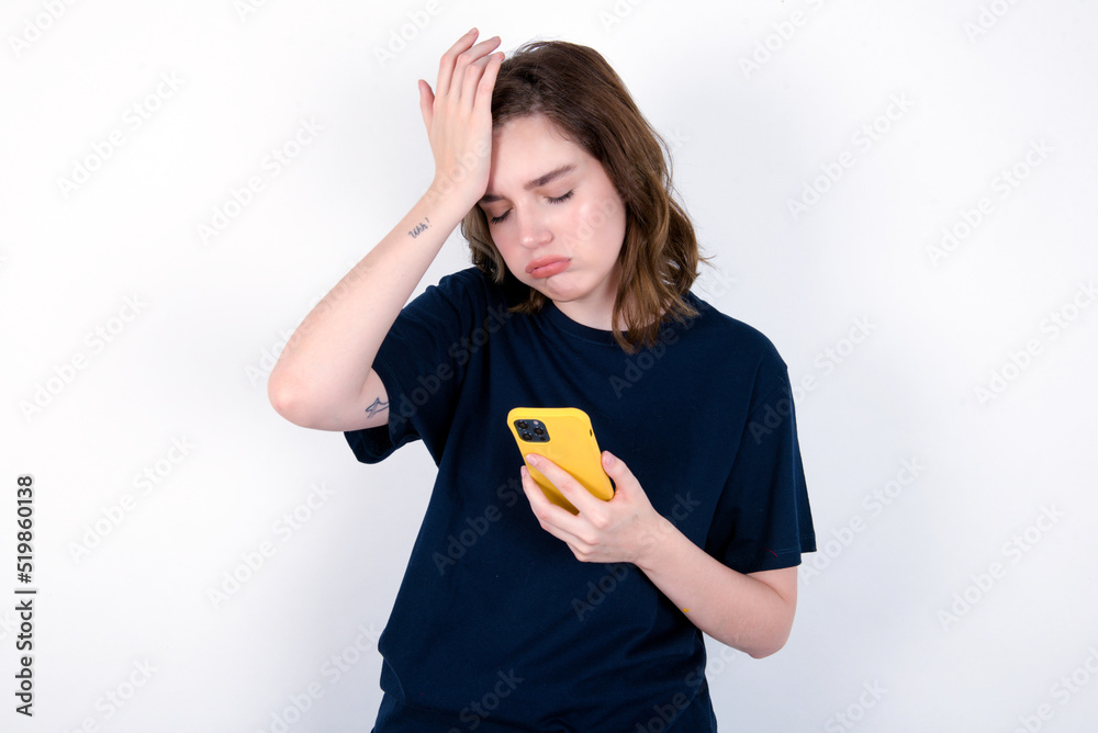 Upset depressed young caucasian woman wearing black T-shirt over white background makes face palm as forgot about something important holds mobile phone expresses sorrow and regret blames Upset depressed young caucasian woman wearing black T-shirt over white background makes face palm as forgot about something important holds mobile phone expresses sorrow and regret blames