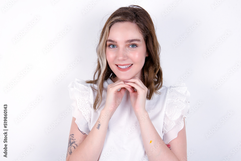 Satisfied young caucasian woman wearing white T-shirt over white background touches chin with both hands, smiles pleasantly, rejoices good day with lover Satisfied young caucasian woman wearing white T-shirt over white background touches chin with both hands, smiles pleasantly, rejoices good day with lover