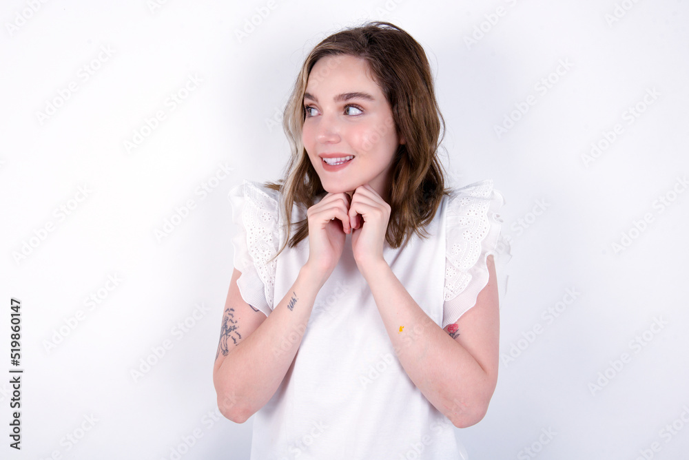 Happy young caucasian woman wearing white T-shirt over white background anticipates something awesome happen, looks happily aside, keeps hands together near face, has glad expression. Happy young caucasian woman wearing white T-shirt over white background anticipates something awesome happen, looks happily aside, keeps hands together near face, has glad expression.