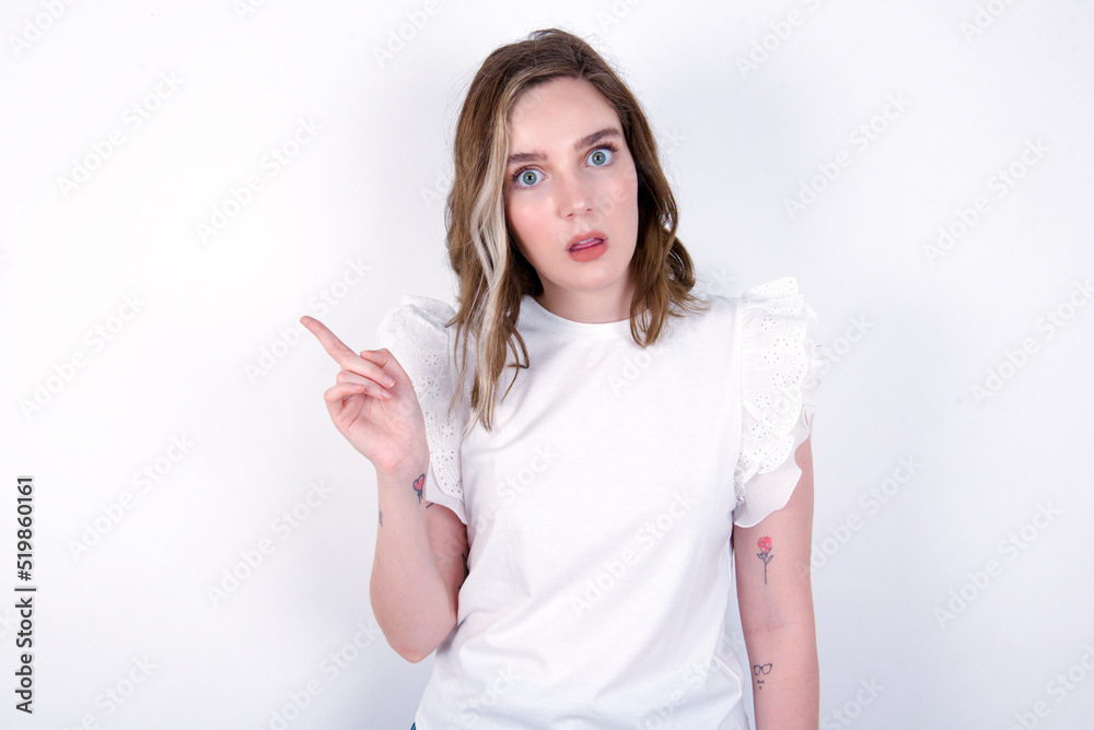 Emotive young caucasian woman wearing white T-shirt over white wall keeps jaw dropped from shock demonstrates amazing promo points right on blank space demonstrates big shopping sale. Advertisement Emotive young caucasian woman wearing white T-shirt over white wall keeps jaw dropped from shock demonstrates amazing promo points right on blank space demonstrates big shopping sale. Advertisement