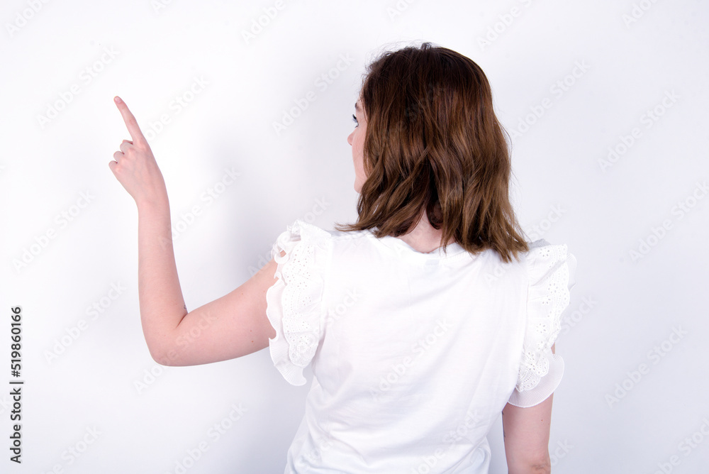young caucasian woman wearing white T-shirt over white background pointing to object on copy space, rear view. Turn your back young caucasian woman wearing white T-shirt over white background pointing to object on copy space, rear view. Turn your back