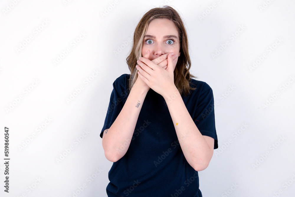 Stunned young caucasian woman wearing black T-shirt over white background covers both hands on mouth, afraids of something astonishing Stunned young caucasian woman wearing black T-shirt over white background covers both hands on mouth, afraids of something astonishing