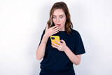 young caucasian woman wearing black T-shirt over white wall being deeply surprised, stares at smartphone display, reads shocking news on website, Omg, its horrible!