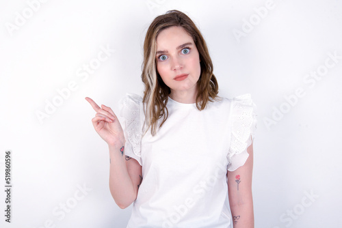 Serious young caucasian woman wearing white T-shirt over white background smirks face points away on copy space shows something unpleasant. Look at this advertisement. Big price concept