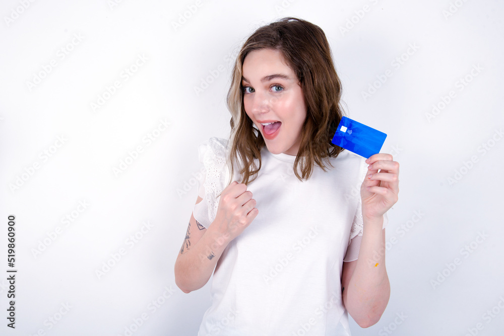 Excited happy positive cheerful smiling young caucasian woman wearing white T-shirt over white background hold credit card raise fist in victory Excited happy positive cheerful smiling young caucasian woman wearing white T-shirt over white background hold credit card raise fist in victory