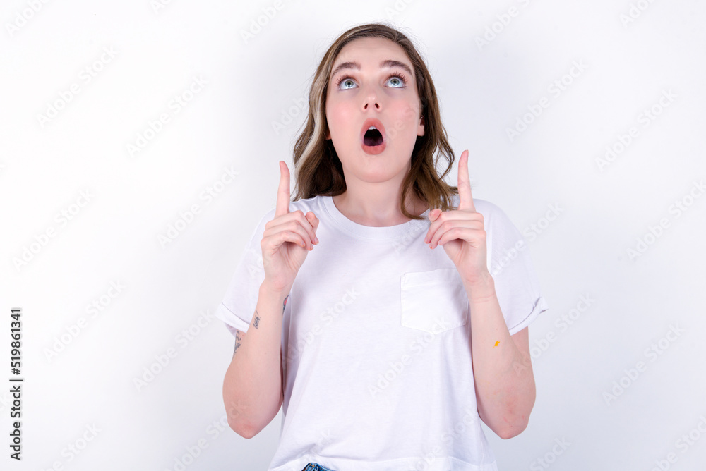 young caucasian woman wearing white T-shirt over white background being amazed and surprised looking and pointing up with fingers showing something strange. young caucasian woman wearing white T-shirt over white background being amazed and surprised looking and pointing up with fingers showing something strange.