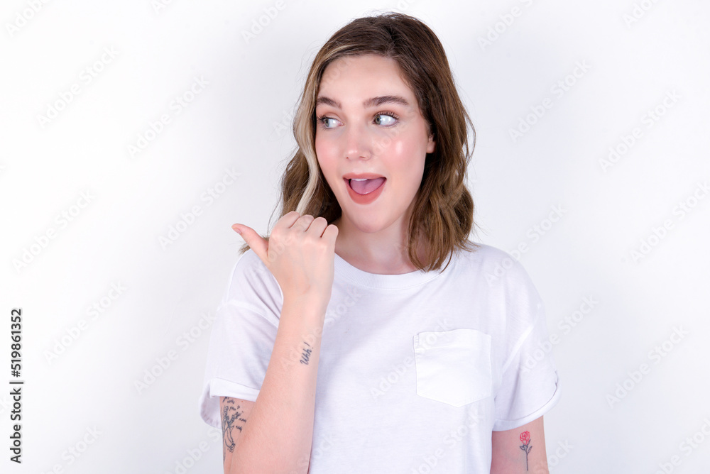 Stupefied young caucasian woman wearing white T-shirt over white wall with surprised expression, opens eyes and mouth widely, points aside with thumb, shows something strange. Advertisement concept. Stupefied young caucasian woman wearing white T-shirt over white wall with surprised expression, opens eyes and mouth widely, points aside with thumb, shows something strange. Advertisement concept.