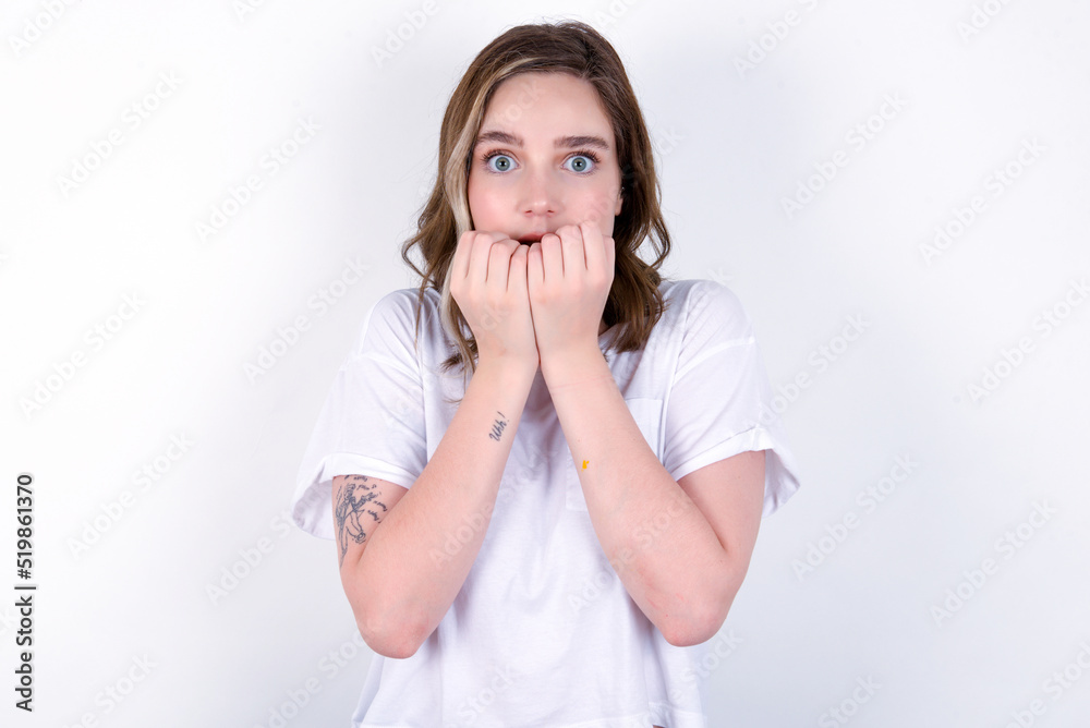 Anxiety - young caucasian woman wearing white T-shirt over white background covering his mouth with hands scared from something or someone bitting nails. Anxiety - young caucasian woman wearing white T-shirt over white background covering his mouth with hands scared from something or someone bitting nails.