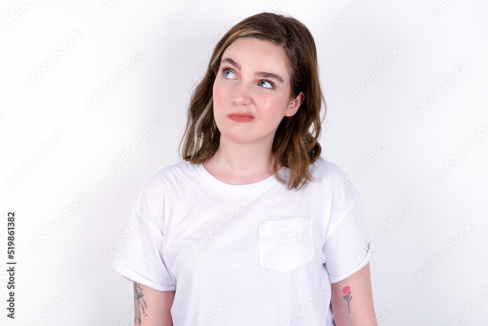 young caucasian woman wearing white T-shirt over white background has worried face looking up lips together, being upset thinking about something important, keeps hands down. young caucasian woman wearing white T-shirt over white background has worried face looking up lips together, being upset thinking about something important, keeps hands down.