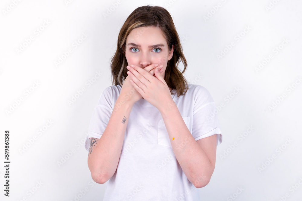 Upset young caucasian woman wearing white T-shirt over white background , covering her mouth with both palms to prevent screaming sound, after seeing or hearing something bad.