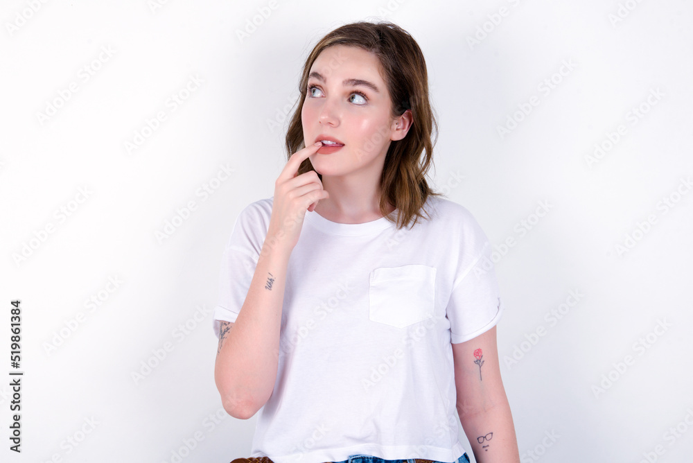 young caucasian woman wearing white T-shirt over white background with thoughtful expression, looks to the camera, keeps hand near face, bitting a finger thinks about something pleasant. young caucasian woman wearing white T-shirt over white background with thoughtful expression, looks to the camera, keeps hand near face, bitting a finger thinks about something pleasant.