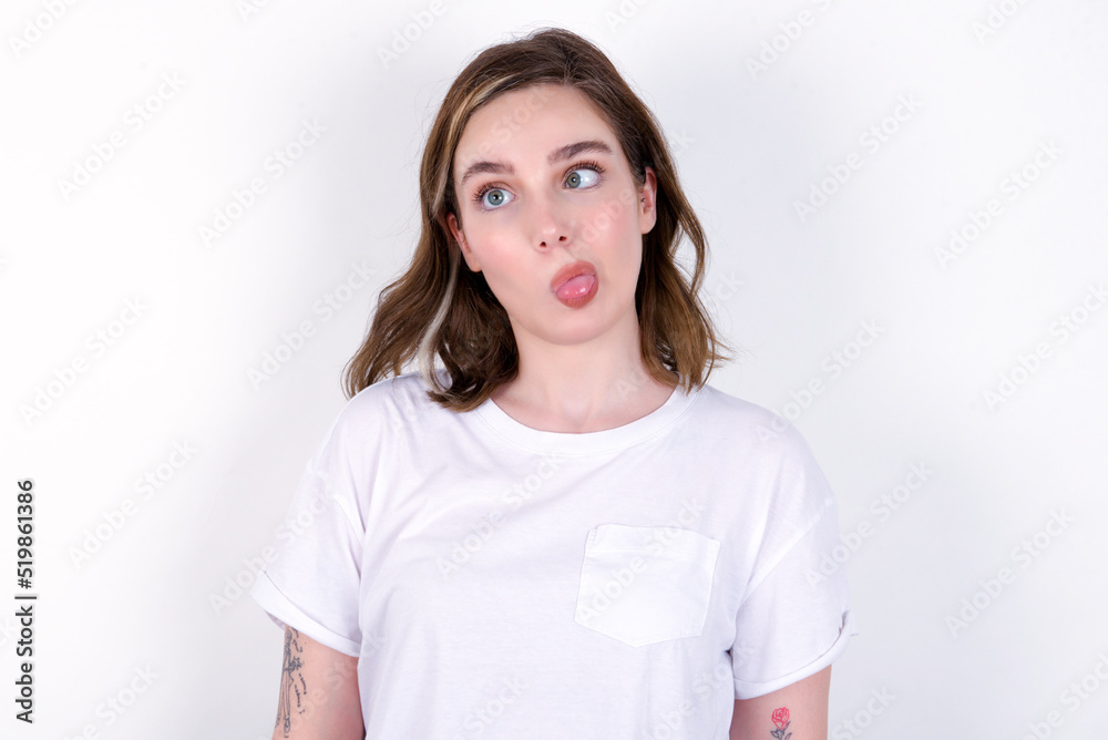 young caucasian woman wearing white T-shirt over white background crosses eyes, puts lips, makes grimace with awkward expression has fun alone, plays fool.