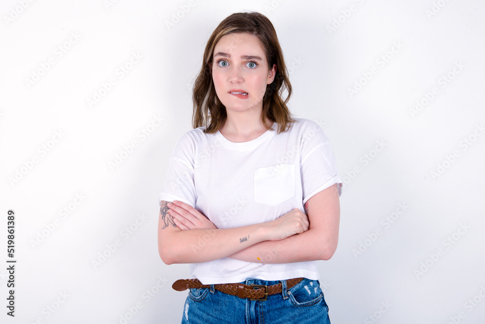 young caucasian woman wearing white T-shirt over white background bitting his mouth and looking worried and scared crossing arms, worry and doubt. young caucasian woman wearing white T-shirt over white background bitting his mouth and looking worried and scared crossing arms, worry and doubt.