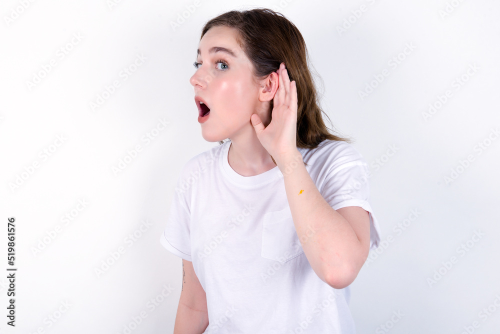 Oh my God! Funny astonished young caucasian woman wearing white T-shirt over white background opening mouth widely looking aside, with hand near ear trying to listen to gossips. Oh my God! Funny astonished young caucasian woman wearing white T-shirt over white background opening mouth widely looking aside, with hand near ear trying to listen to gossips.