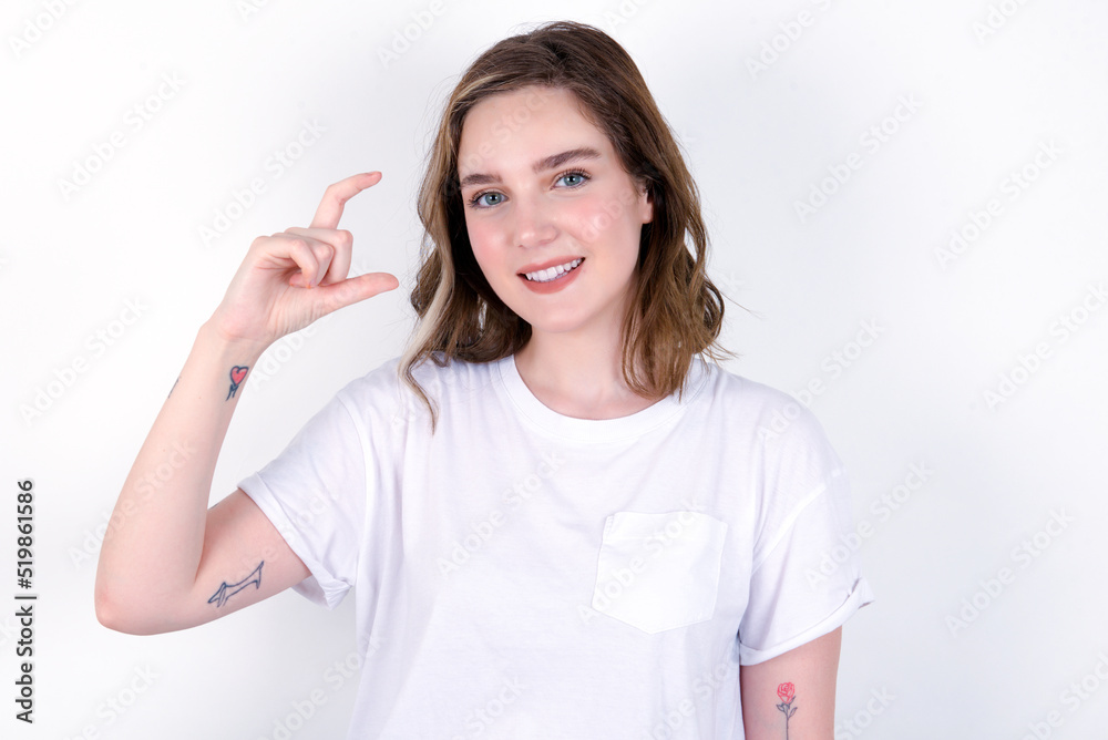 young caucasian woman wearing white T-shirt over white background smiling and gesturing with hand small size, measure symbol. young caucasian woman wearing white T-shirt over white background smiling and gesturing with hand small size, measure symbol.