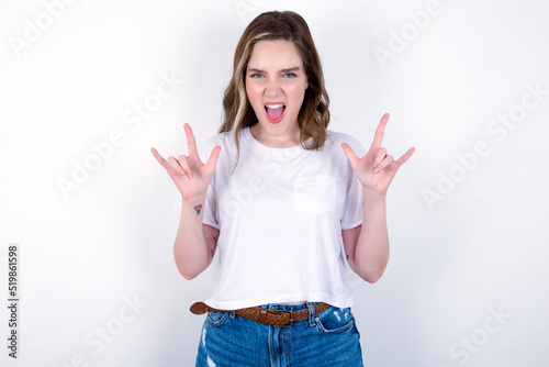 Born to rock this world. Joyful young caucasian woman wearing white T-shirt over white background screaming out loud and showing with raised arms horns or rock gesture.