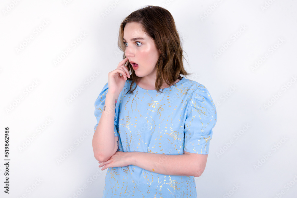 Astonished young caucasian woman wearing blue T-shirt over white background looks aside surprisingly with opened mouth. Astonished young caucasian woman wearing blue T-shirt over white background looks aside surprisingly with opened mouth.