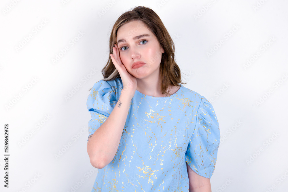 Sad lonely young caucasian woman wearing blue T-shirt over white background touches cheek with hand bites lower lip and gazes with displeasure. Bad emotions Sad lonely young caucasian woman wearing blue T-shirt over white background touches cheek with hand bites lower lip and gazes with displeasure. Bad emotions
