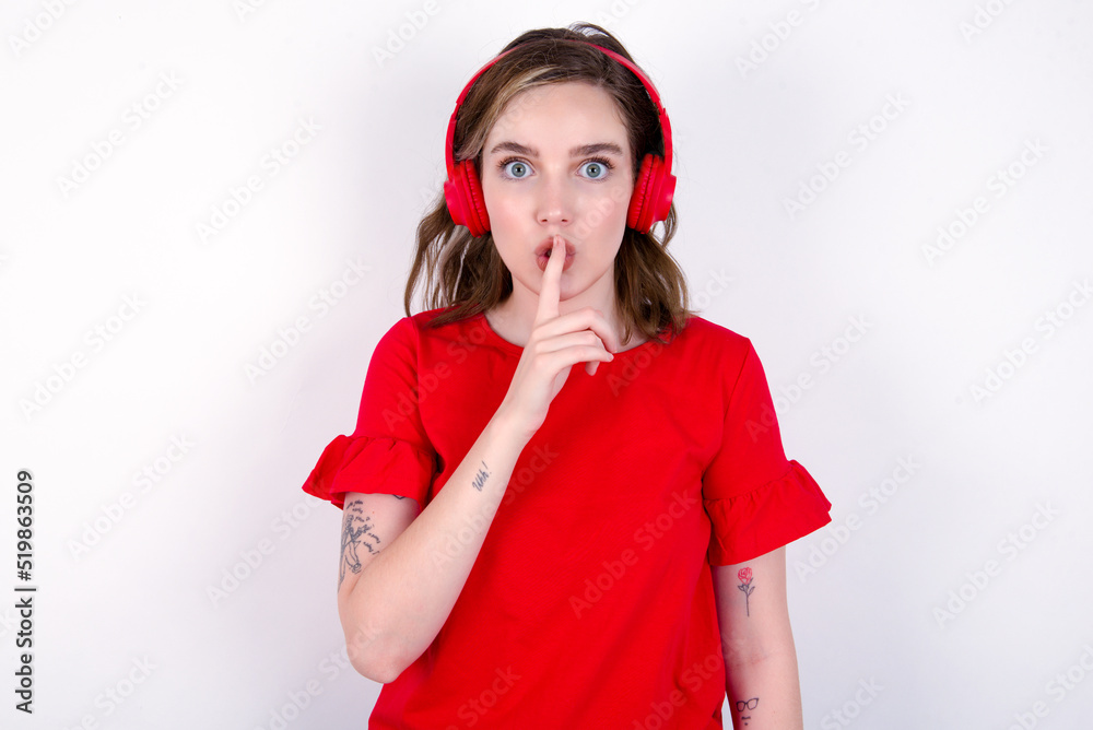 young caucasian woman wearing red T-shirt over white background making hush gesture with finger on her lips wearing wireless headphones. Be quiet. young caucasian woman wearing red T-shirt over white background making hush gesture with finger on her lips wearing wireless headphones. Be quiet.