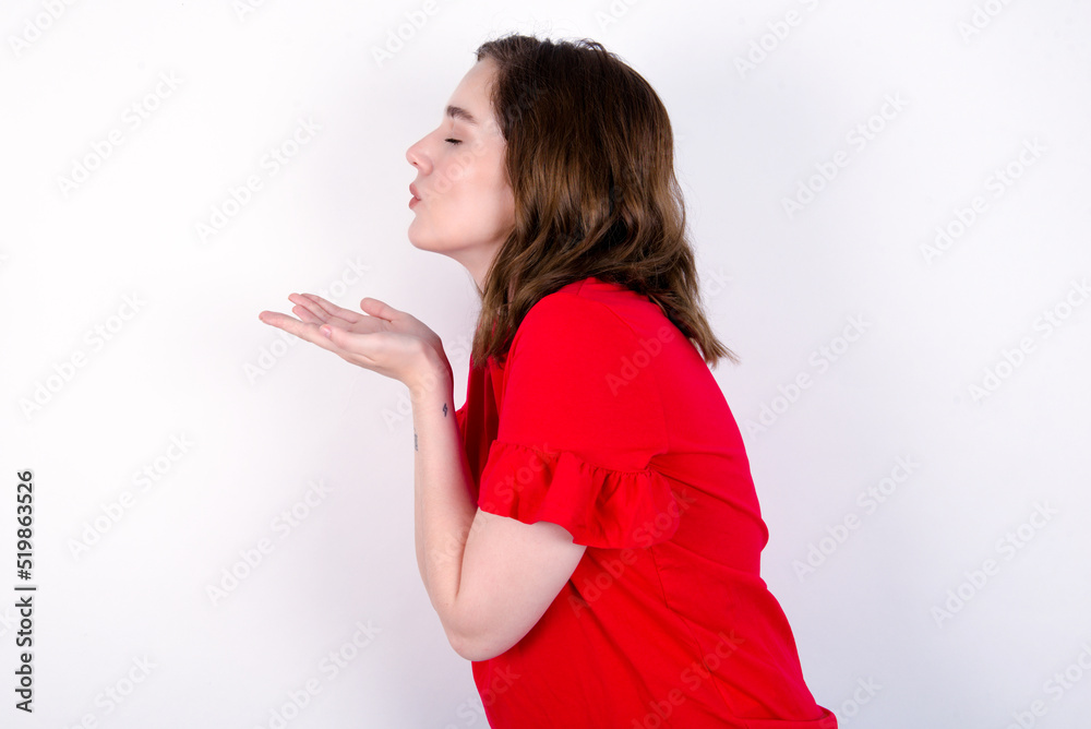 Profile side view view portrait of attractive young caucasian woman wearing red T-shirt over white background sending air kiss Profile side view view portrait of attractive young caucasian woman wearing red T-shirt over white background sending air kiss