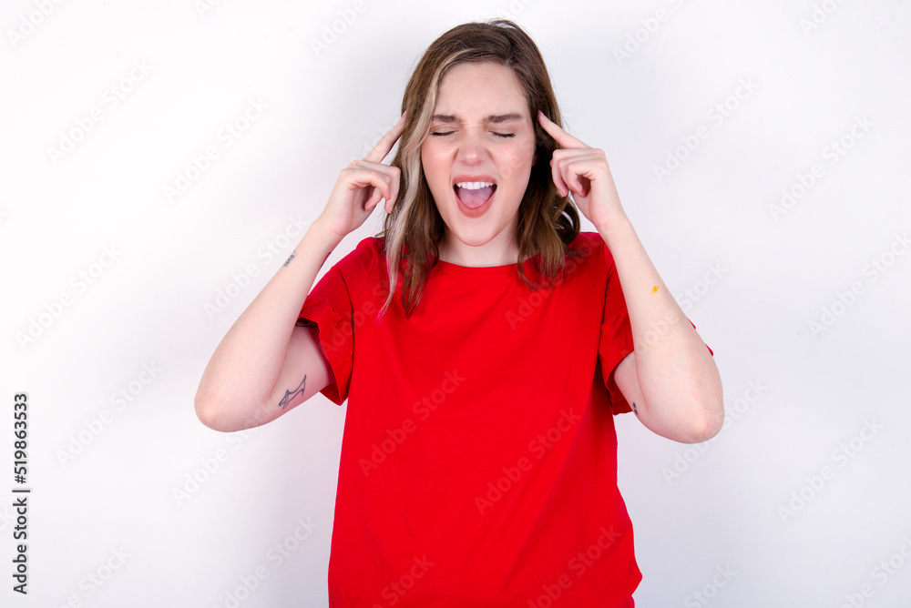 Photo of crazy young caucasian woman wearing red T-shirt over white background screaming and pointing with fingers at hair closed eyes Photo of crazy young caucasian woman wearing red T-shirt over white background screaming and pointing with fingers at hair closed eyes