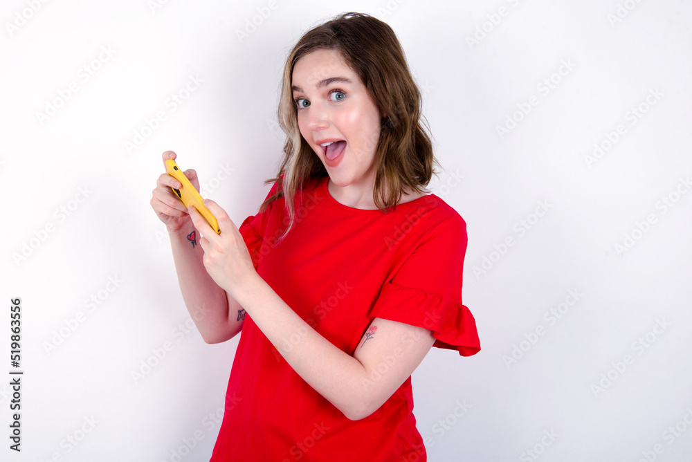 Nice addicted cheerful young caucasian woman wearing red T-shirt over white background using gadget playing network game Nice addicted cheerful young caucasian woman wearing red T-shirt over white background using gadget playing network game