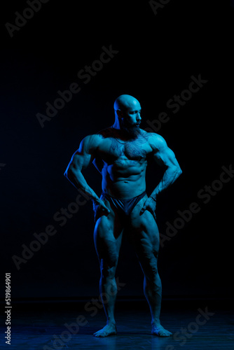 Athletic man demonstrates muscles in the light of a blue light filter on a dark background.
