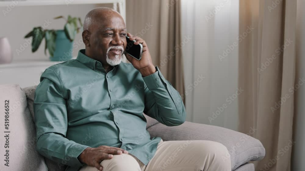 African bald middle-aged man senior mature businessman sitting on sofa at home in office talking phone chatting mobile conversation making order with smartphone discussing remote talking sharing news