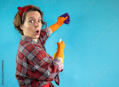Cheerful adult woman in a plaid shirt wipes with a purple rag on a blue isolated background. Woman pin up cleaning holding a spray bottle in her hand