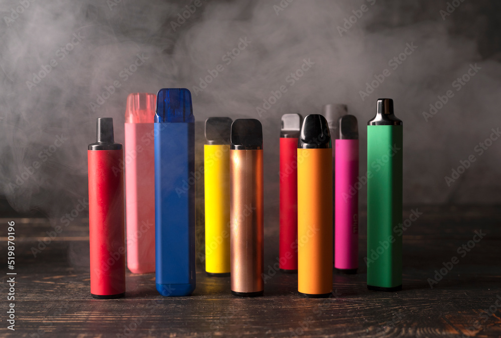Set of colorful disposable electronic cigarettes on a dark wood background with smoke. - obrazy, fototapety, plakaty 