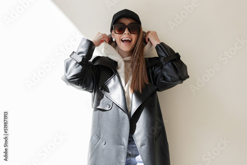 Happiness young beautiful fashion woman hipster with happy emotions with trendy sunglasses and black hat in leather coat and hoodie wears a hood have fun enjoy near a wall on the street in sunlight