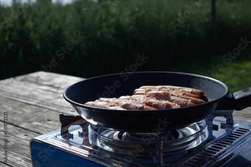 Tableau sur toile Sausages wrapped in bacon are fried in a frying pan on a gas camping stove again